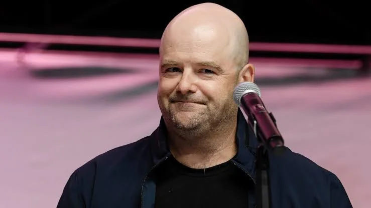 Smiling Bald Man Wearing a Dark Jacket Stands at a Microphone on Stage with Pink Lighting in the Background